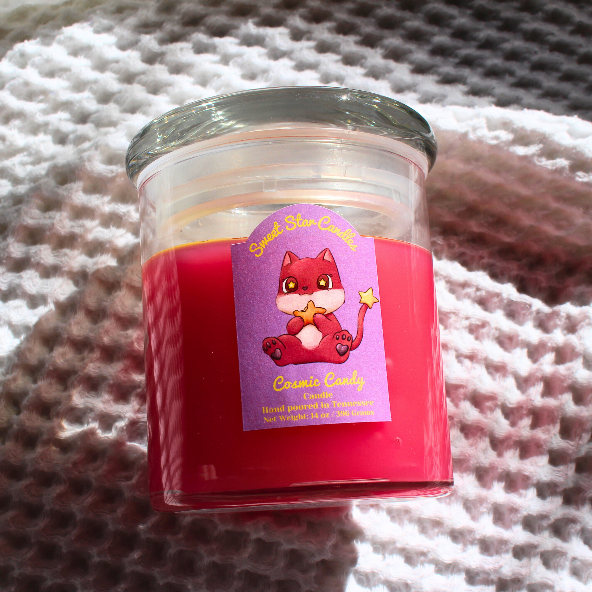 A magenta candle with glass lid with yellow stars sitting on top of the candle the label features a magenta creature with a white chin and belly with cute stars in his eyes munching on a orange star cosmic candy he also has a tail with a star on the end the label reads sweet star candles cosmic candy candle hand poured in Tennessee with a net weight of 14 ounces