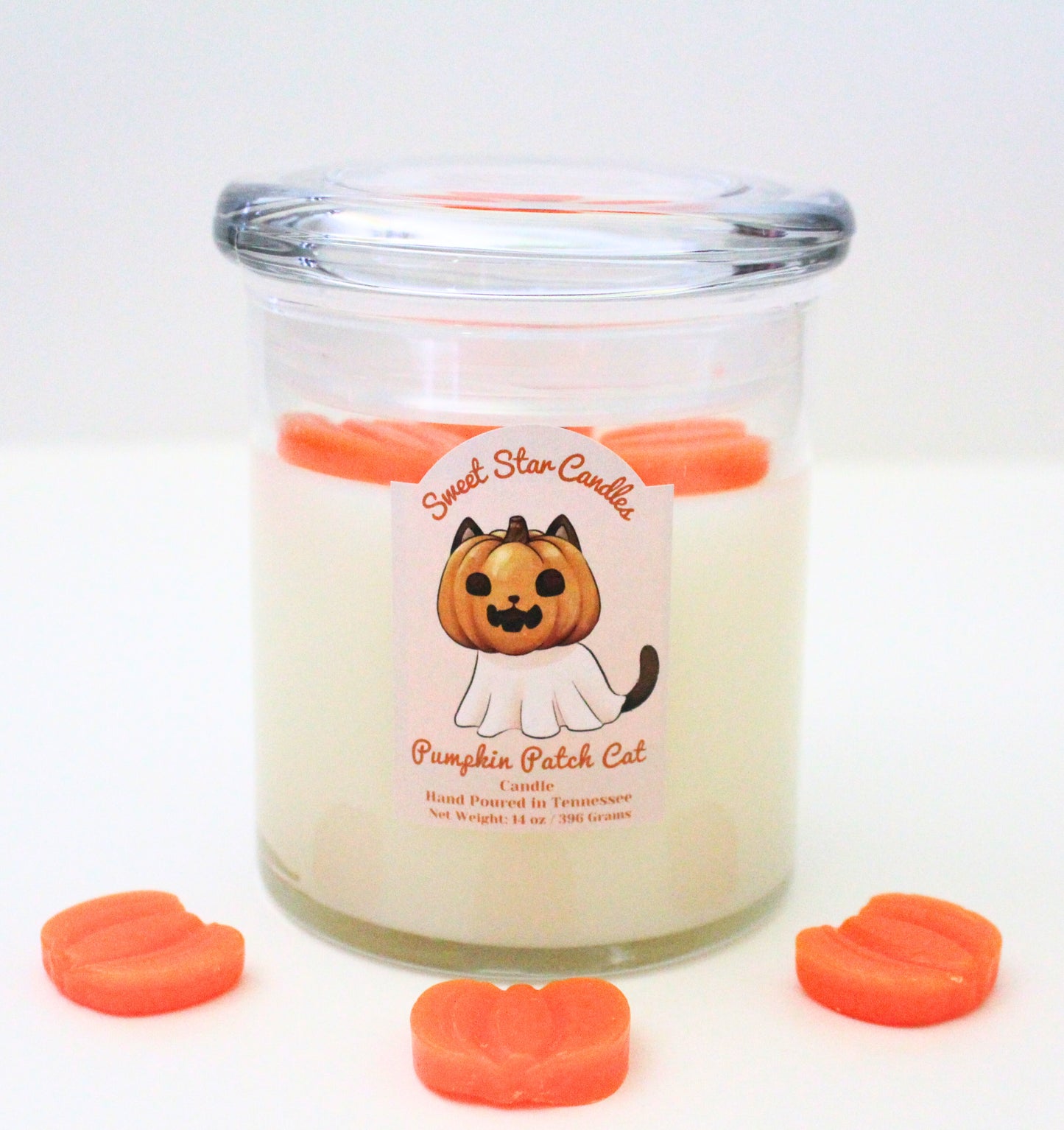 our pumpkin patch cat candle has white wax on the bottom with pumpkin embeds on top and surrounding it on the ground the label features a cat with a jack o lantern face smiling with a ghost sheet his ears poke out of the pumpkin and tail out of the cloth the label reads pumpkin patch cat the scent name under that hand poured in east Tennessee weight 14 oz or 150 grams