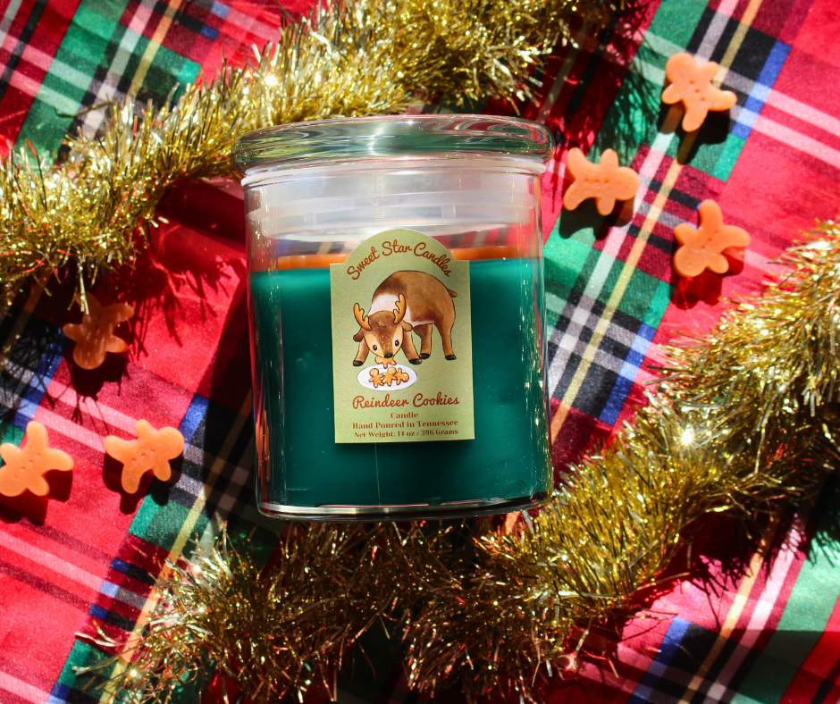 our reindeer cookies candle sits over a festive background our candle is a glass jar and glass lid the label reads sweet star candles on top in light brown cursive letters under that a reindeer eats gingerbread cookies from a plate intended for santa the reindeer is in a cute style under that it reads reindeer cookies the candle scent under that reads i sparkle as i melt hand poured in Tennessee weight 14 oz or 150 grams