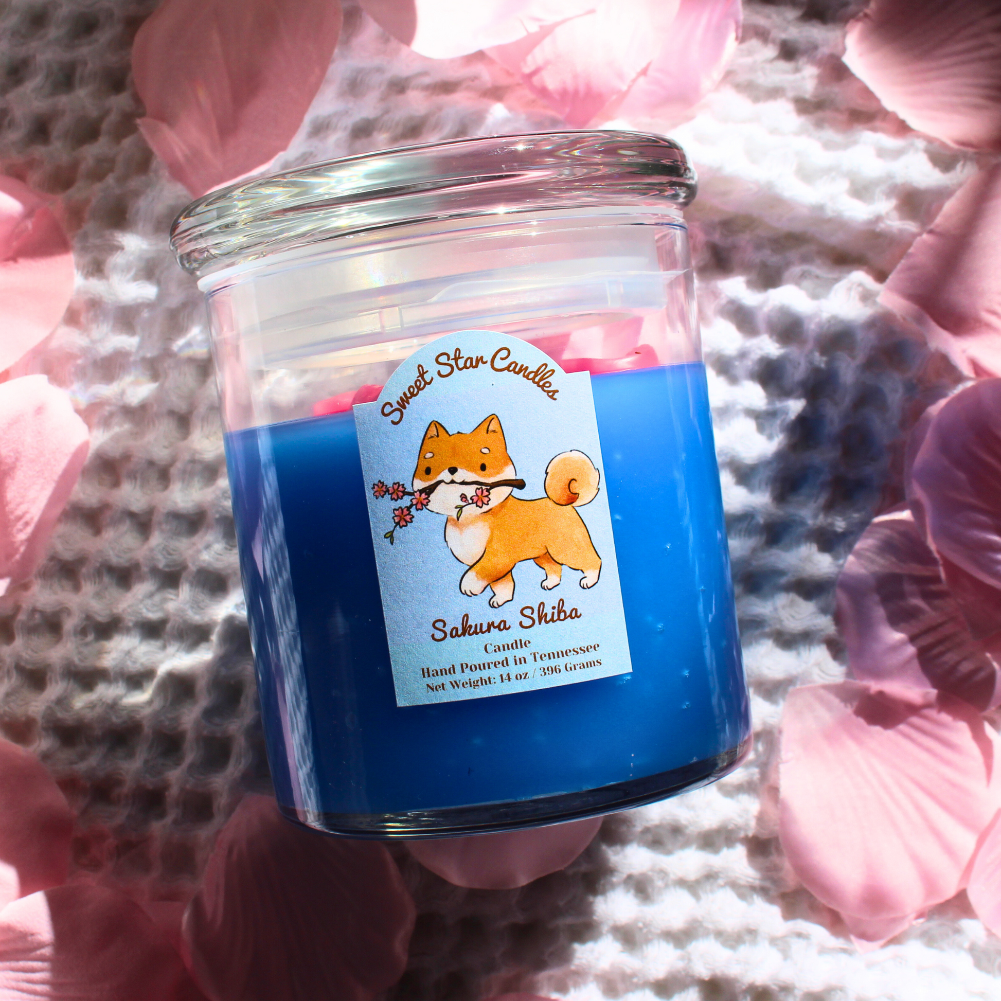 This candle has a light blue base with magenta flower petals sitting on top the label features a shiba inu dog prancing with a cherry blossom branch in his mouth the label reads sweet star candles sakura shiba candle hand poured in tennessee with a net weight of 14 ounces