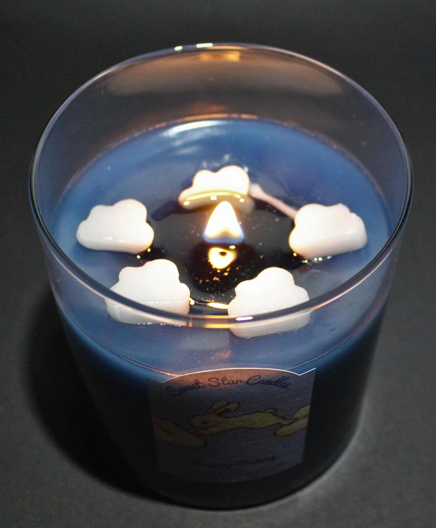 the breezy bunny candle candle is lit and small sparkles and glitter can be seen around the flame in the melted wax the clouds are slowly melting into the dark blue wax