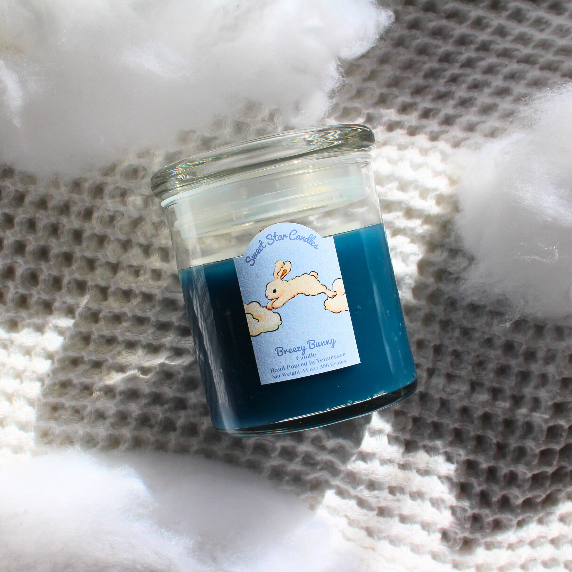 our breezy bunny sparkle as it melts candle sits on a white background with clouds the label reads sweet star candles with a bunny underneath jumping from cloud to cloud under that it says breezy bunny candle hand poured in tennessee net weight 14 ounces the label is light blue with a white bunny the wax is dark blue in a glass vessel