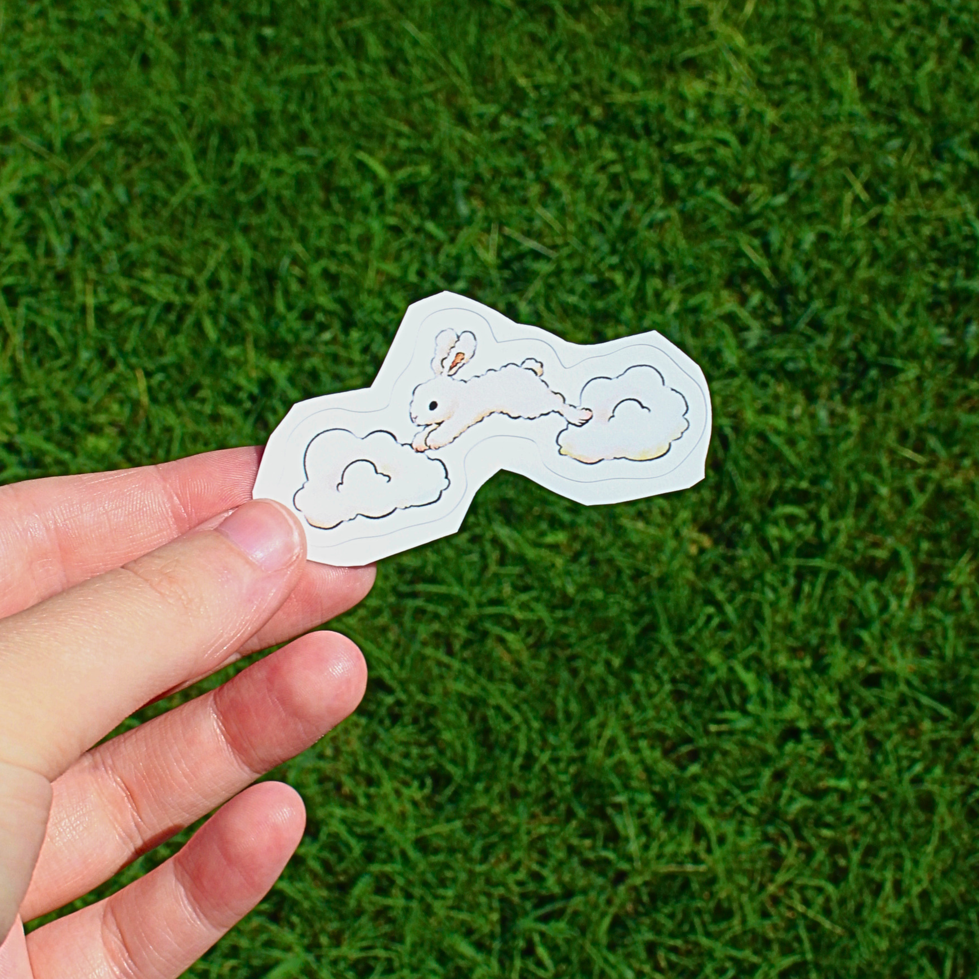 A hand holding a small white sticker with a bunny and cloud illustration against a green grass background.