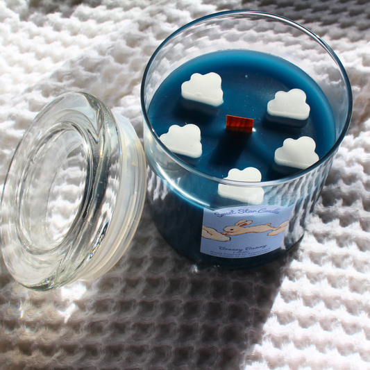 this is a top view of our breezy bunny candle you can see white cloud embeds surrounding a wooden wick the wax is dark blue in a glass container vessel