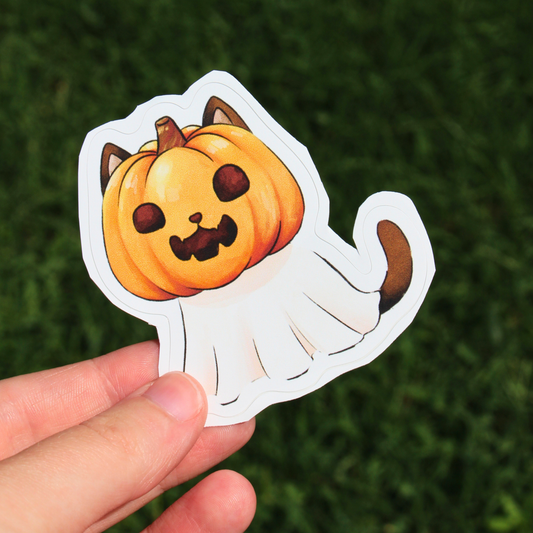 A hand holding a die-cut sticker of a cute cartoon pumpkin cat ghost, set against a blurred green grass background.