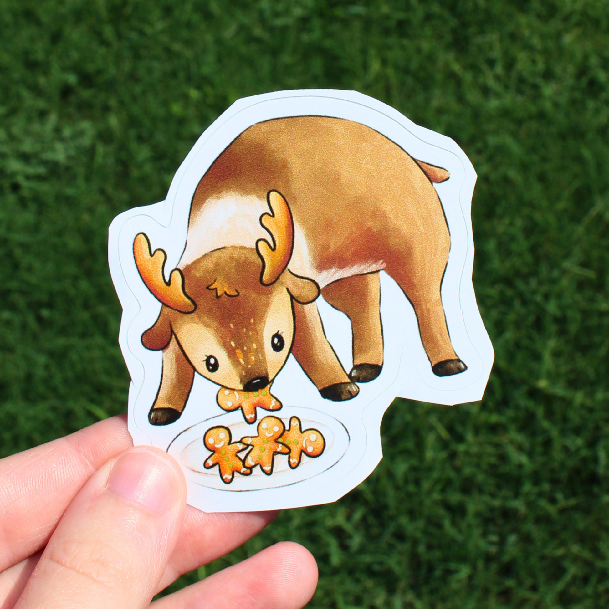 A hand holds a die-cut sticker featuring a cute cartoon reindeer eating gingerbread cookies, set against a blurred green grass background.