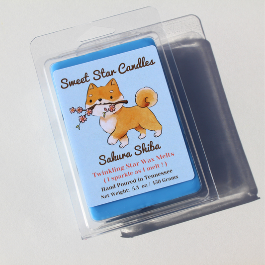 our sakura shiba wax melt sits on a white background the soy wax is a light blue color on top of the clamshell a lebel reads sweet star candles on top in a dark brown color under that the shiba inu dog marches proudly holding a cherry blossom stick in his mouth he is a typical shiba inu and the branch has small pink flowers on the stems under that reads sakura shiba the name of the scent under that is says i sparkle as i melt hand poured in east Tennessee 5.3 ounces or 150 grams