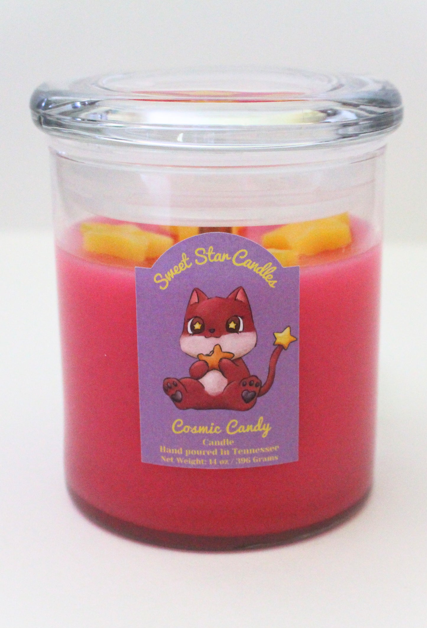 A magenta candle with glass lid with yellow stars sitting on top of the candle the label features a magenta creature with a white chin and belly with cute stars in his eyes munching on a orange star cosmic candy he also has a tail with a star on the end the label reads sweet star candles cosmic candy candle hand poured in Tennessee with a net weight of 14 ounces