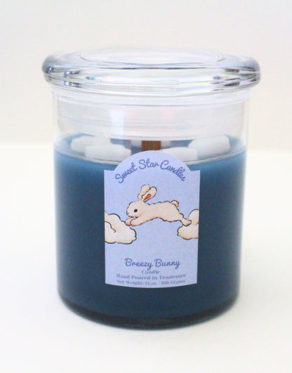 Dark blue candle with white clouds, wooden wick, and a cute label with a white rabbit jumping between two clouds the label reads sweet star candles breezy bunny candle hand poured in tennessee with a net weight of 14 ounces