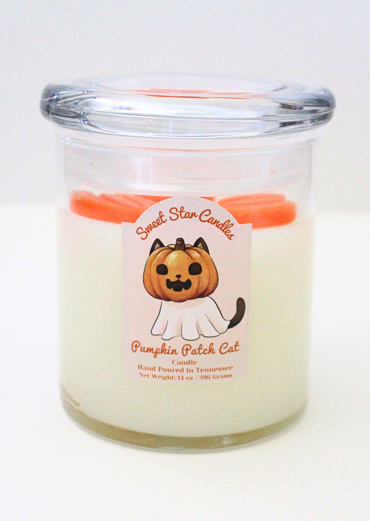 The candle feautures a white base with orange pumpkins on top of the wax with a glass lid the label features a black cat with a pumpkin on his head and a sheet ressembling a ghost covering the rest of his body his face also features a wide grin and his tail pokes out from beneath the sheet the label also reads sweet star candles pumpkin patch cat candle hand poured in tennessee with a net weight of 14 ounces