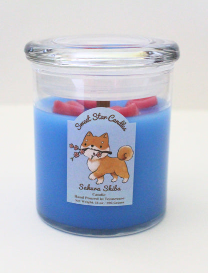 This candle has a light blue base with magenta flower petals sitting on top the label features a shiba inu dog prancing with a cherry blossom branch in his mouth the label reads sweet star candles sakura shiba candle hand poured in tennessee with a net weight of 14 ounces